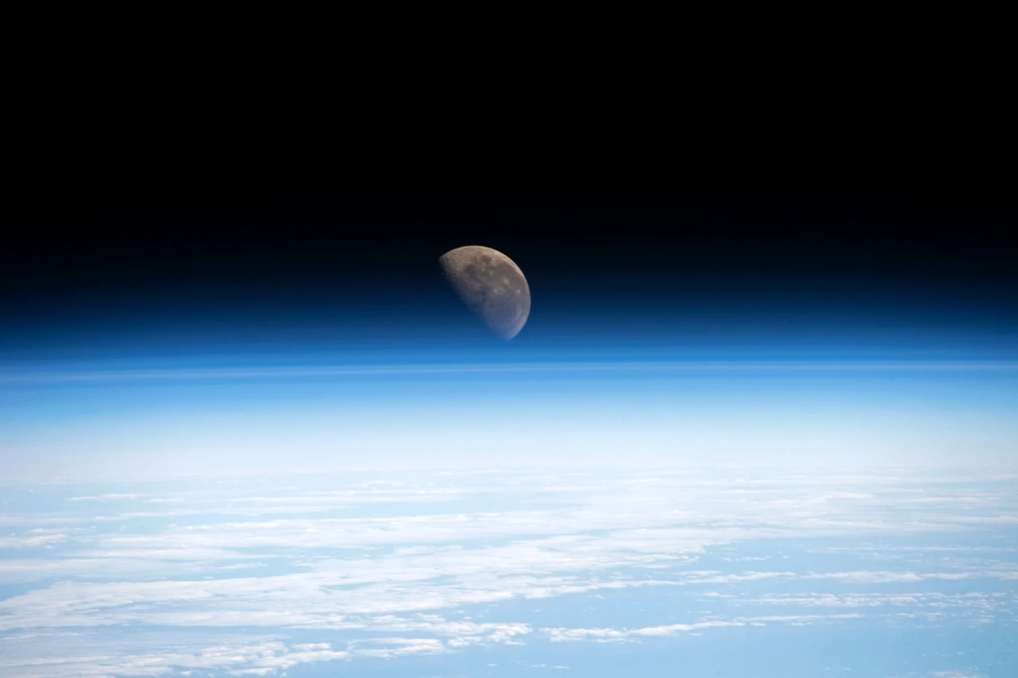NASA宇航员拍摄的地球大气侧影与远处月球，低处云层呈浅蓝色，高处渐入黑暗